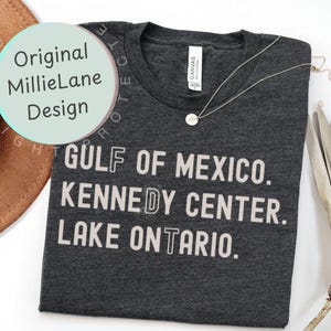 May include: A dark charcoal heather gray t-shirt with a crew neckline and a white graphic print that reads GULF OF MEXICO. KENNEDY CENTER. LAKE ONTARIO. The shirt features a white brand label at the inner neck and a thin silver chain necklace.