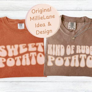 Op de afbeelding: Twee t-shirts met retro tekstontwerpen. Eén is verbrand oranje met "SWEET POTATO" in lichtoranje. De andere is bruin met "KIND OF RUDE POTATO" in lichtoranje. Een zilveren ketting met een hanger ligt op het bruine shirt.