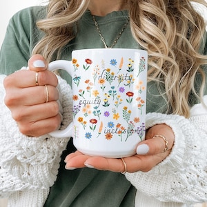 May include: White ceramic mug with a floral pattern and the words "diversity", "equity", and "inclusion" written in black.