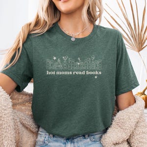 Op de afbeelding: Een donkergroen gemêleerd T-shirt met korte mouwen en een witte lijntekening van een rij boeken, vazen en bloemen. De tekst "hot moms read books" is in een wit schreeflettertype onder de illustratie gedrukt, geaccentueerd door kleine sterretjes.
