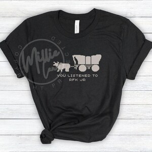 Puede incluir: Camiseta negra con una imagen pixelada de un oso tirando de una carreta cubierta. El texto "YOU LISTENED TO RFK JR" est&aacute; debajo. La camiseta est&aacute; hecha de un material suave y tiene cuello redondo.