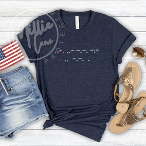 May include: Navy blue t-shirt with a Morse code design. The shirt is paired with denim shorts, sandals, sunglasses, and a small American flag. The shirt has a tag that says "Canvas".