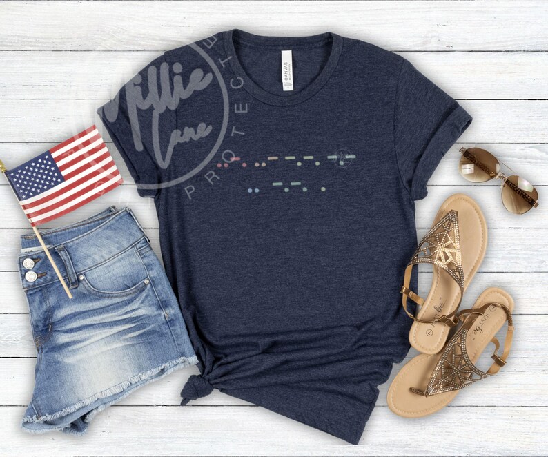 May include: Navy blue t-shirt with a Morse code design. The shirt is paired with denim shorts, sandals, sunglasses, and a small American flag. The shirt has a tag that says "Canvas".