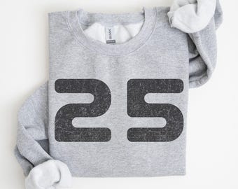 以下が含まれることがあります： 大きな、使い古された黒いフォントで「25」の数字がプリントされた、明るい灰色のスウェットシャツ。スウェットシャツはきれいに折りたたまれ、袖と襟の内側が見えます。生地は柔らかく快適に見えます。