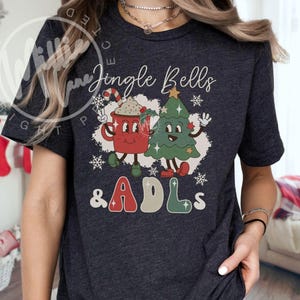 May include: A dark heather gray short-sleeve t-shirt featuring a graphic print of a red mug with a candy cane and a green Christmas tree with faces. The text reads Jingle Bells & ADLs in a retro font, surrounded by white snowflakes and stars.