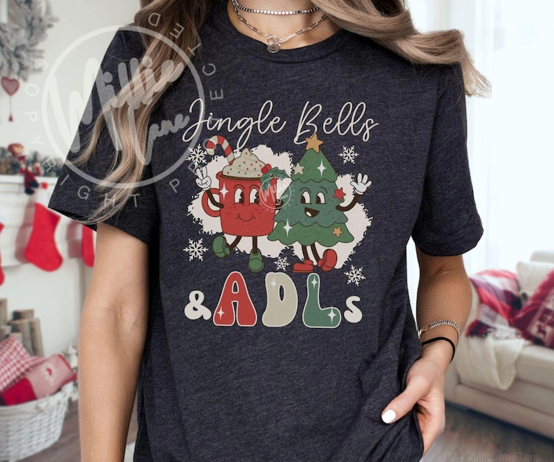 May include: A dark heather gray short-sleeve t-shirt featuring a graphic print of a red mug with a candy cane and a green Christmas tree with faces. The text reads Jingle Bells & ADLs in a retro font, surrounded by white snowflakes and stars.