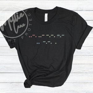 May include: Black t-shirt with a Morse code design in pastel colors. The shirt has a crew neck and short sleeves. The image also includes the text "Millie Lane" in a circular design.