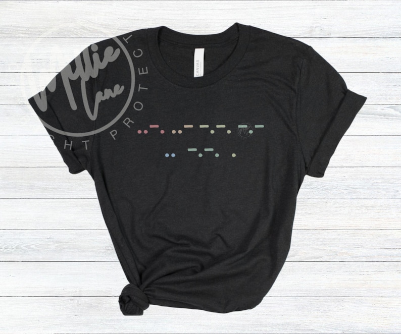 May include: Black t-shirt with a Morse code design in pastel colors. The shirt has a crew neck and short sleeves. The image also includes the text "Millie Lane" in a circular design.