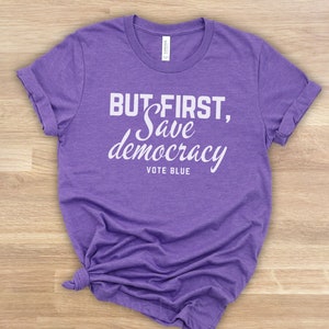 May include: A lilac t-shirt with the white text "BUT FIRST, Save democracy VOTE BLUE". The shirt is made of a soft fabric and has rolled-up sleeves. The shirt is knotted at the bottom.