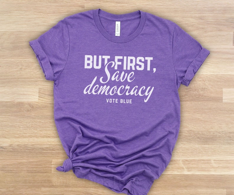 May include: A lilac t-shirt with the white text "BUT FIRST, Save democracy VOTE BLUE". The shirt is made of a soft fabric and has rolled-up sleeves. The shirt is knotted at the bottom.