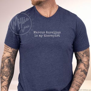 以下が含まれることがあります： 白文字で「Marcus Aurelius is my therapist」と書かれたネイビーブルーのVネックTシャツ。柔らかい素材で作られています。カジュアルウェアです。