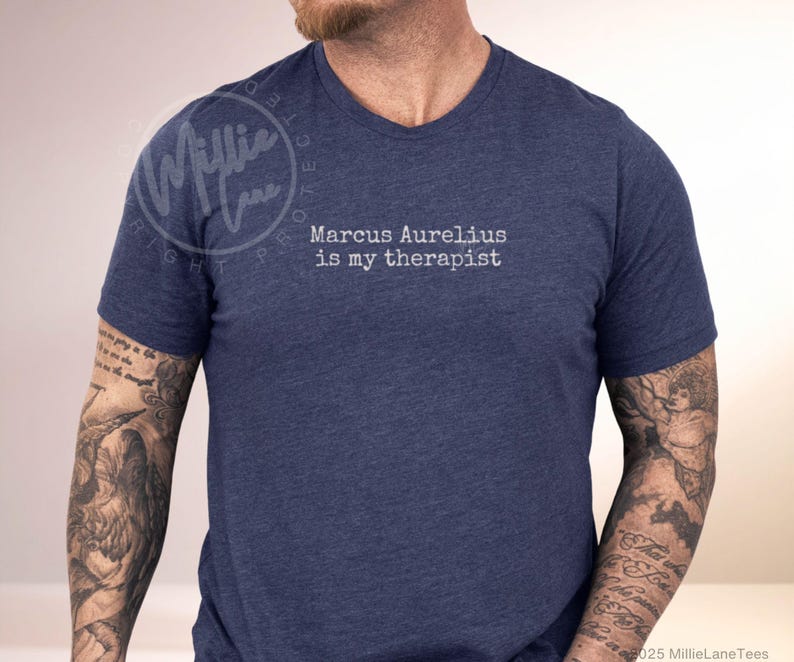 以下が含まれることがあります： 白文字で「Marcus Aurelius is my therapist」と書かれたネイビーブルーのVネックTシャツ。柔らかい素材で作られています。カジュアルウェアです。