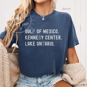 May include: A dark blue short-sleeve t-shirt featuring white text that reads GULF OF MEXICO. KENNEDY CENTER. LAKE ONTARIO. The shirt has a crew neckline and a relaxed fit, paired with light blue denim shorts and a silver chain necklace.
