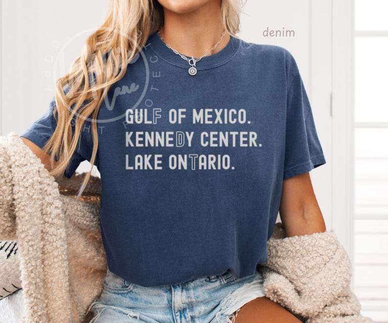 May include: A dark blue short-sleeve t-shirt featuring white text that reads GULF OF MEXICO. KENNEDY CENTER. LAKE ONTARIO. The shirt has a crew neckline and a relaxed fit, paired with light blue denim shorts and a silver chain necklace.
