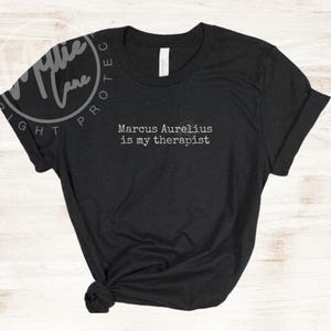 以下が含まれることがあります： 「Marcus Aurelius is my therapist」という白文字がプリントされた黒いTシャツ。クルーネックで袖は折り返され、裾は結ばれています。カジュアルな印象です。