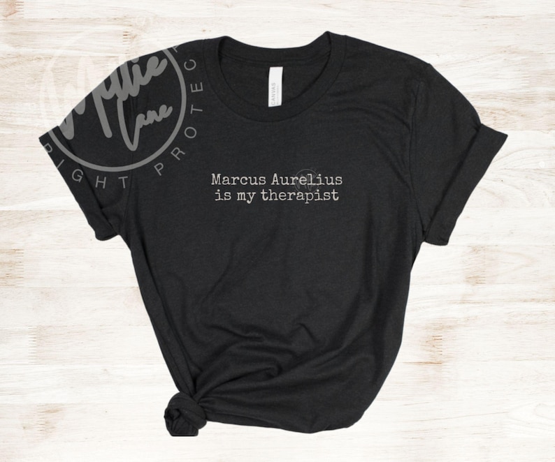 以下が含まれることがあります： 「Marcus Aurelius is my therapist」という白文字がプリントされた黒いTシャツ。クルーネックで袖は折り返され、裾は結ばれています。カジュアルな印象です。