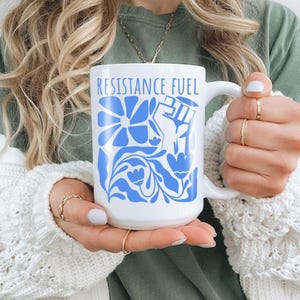 May include: White ceramic mug featuring the words "RESISTANCE FUEL" in blue, accompanied by a blue floral design and a raised fist. The mug has a large handle and is held by a person wearing a white sweater.