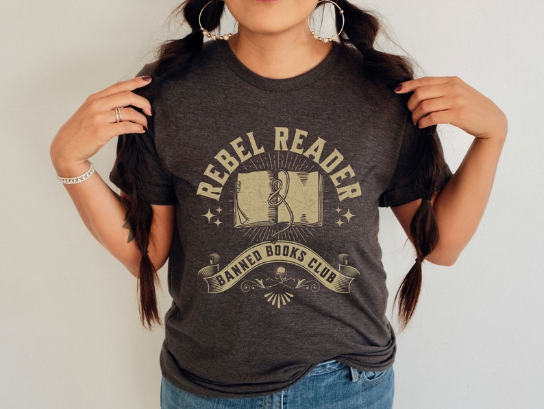 K&ouml;nnte beinhalten: Ein dunkelgraues T-Shirt mit einem Vintage-Grafikdesign, auf dem "Rebel Reader Banned Books Club" steht. Die Grafik zeigt ein geschlossenes Buch mit einem Lesezeichenband und Sternen.