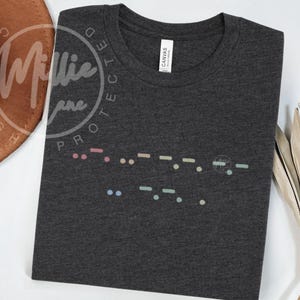 May include: Dark gray t-shirt with a colorful Morse code design. The shirt features a crew neck and short sleeves. The Morse code is in pastel colors and is printed across the chest.