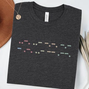 May include: A dark heather grey t-shirt with a colorful Morse code design. The code reads "HOPE".