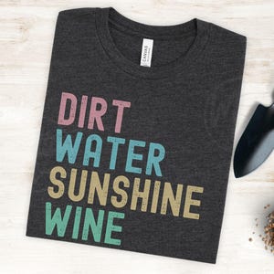 May include: Dark grey t-shirt with the words "DIRT WATER SUNSHINE WINE" in pink, teal, tan, and green block letters. A small gardening trowel and other gardening tools are visible in the background.