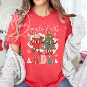 May include: A red short-sleeve t-shirt featuring a graphic print of a smiling red mug and a green Christmas tree with arms and legs. The text reads Jingle Bells & ADLs, surrounded by white snowflake accents and small star shapes.