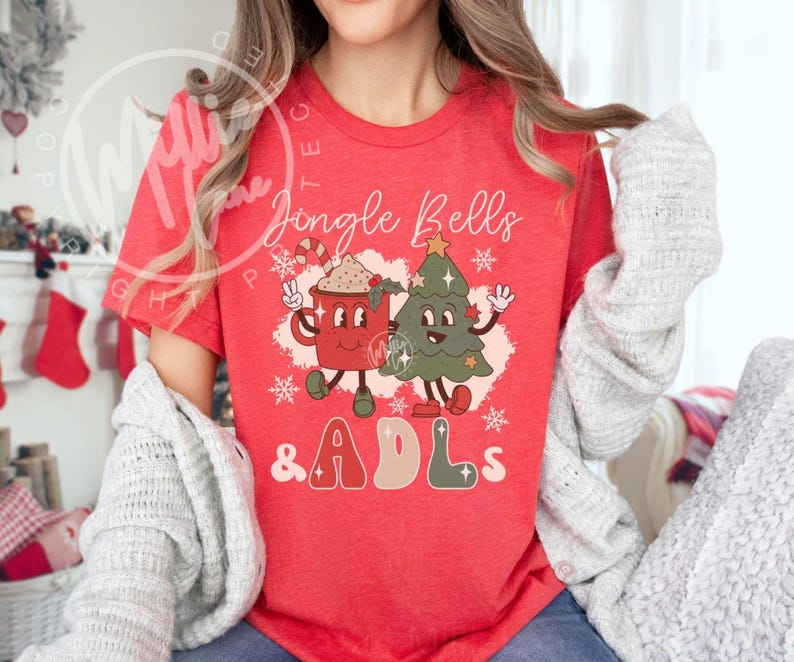 May include: A red short-sleeve t-shirt featuring a graphic print of a smiling red mug and a green Christmas tree with arms and legs. The text reads Jingle Bells & ADLs, surrounded by white snowflake accents and small star shapes.