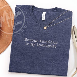 以下が含まれることがあります： 「Marcus Aurelius is my therapist」の白文字が入ったネイビーブルーのTシャツ。丸いペンダントが付いたゴールドカラーのネックレスがシャツの上にあります。シャツは柔らかい素材で作られています。