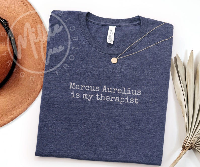 以下が含まれることがあります： 「Marcus Aurelius is my therapist」の白文字が入ったネイビーブルーのTシャツ。丸いペンダントが付いたゴールドカラーのネックレスがシャツの上にあります。シャツは柔らかい素材で作られています。