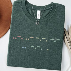 May include: A dark green t-shirt with a colorful Morse code design across the front. The shirt has a crew neck and short sleeves. A silver necklace with a sun pendant is on the shirt. The shirt is made of a soft material.