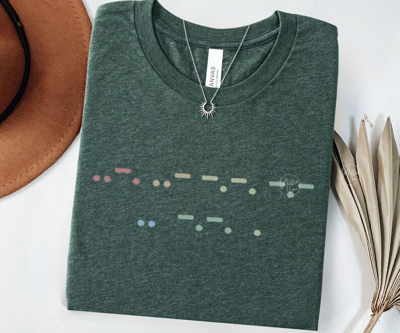 May include: A dark green t-shirt with a colorful Morse code design across the front. The shirt has a crew neck and short sleeves. A silver necklace with a sun pendant is on the shirt. The shirt is made of a soft material.
