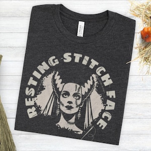 May include: Dark gray t-shirt with the text "RESTING STITCH FACE" in a semi-circle around a Bride of Frankenstein graphic. The graphic is in a vintage style, with a needle and thread. The shirt is on a white wooden surface.