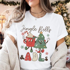 May include: A white short-sleeve t-shirt featuring a graphic print of a red mug with a candy cane and a green Christmas tree with arms and legs. The text Jingle Bells & ADLs is printed in a retro font. The design includes small red and green snowflakes.