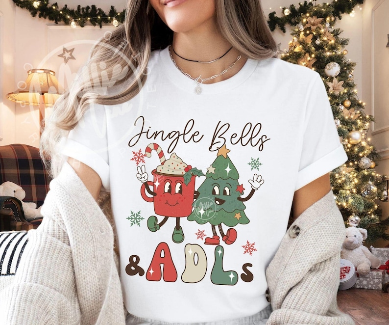 May include: A white short-sleeve t-shirt featuring a graphic print of a red mug with a candy cane and a green Christmas tree with arms and legs. The text Jingle Bells & ADLs is printed in a retro font. The design includes small red and green snowflakes.