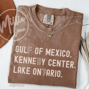 May include: A brown short-sleeved t-shirt with a crew neckline featuring the text GULF OF MEXICO. KENNEDY CENTER. LAKE ONTARIO. printed in a light beige color. The shirt has a visible brand label on the inside collar and a faded, washed-out finish.