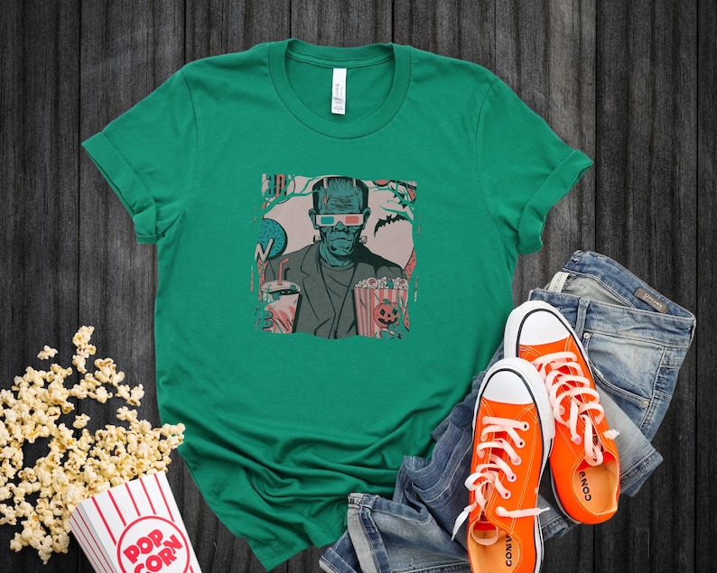 May include: A green t-shirt with a graphic of Frankenstein wearing 3D glasses and holding a bucket of popcorn. The graphic is in a retro comic book style.