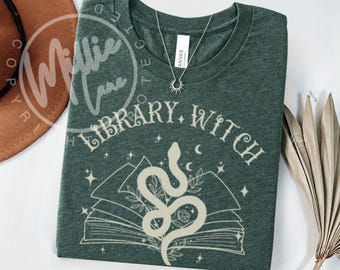 May include: A dark forest green t-shirt featuring a cream-colored graphic of an open book with a snake coiled on top, surrounded by stars and leaves. The text "LIBRARY WITCH" is printed in a decorative font above the book illustration.