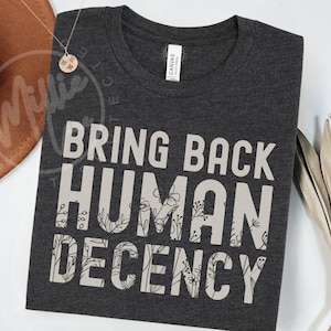 Op de afbeelding: Donkergrijs t-shirt met de tekst "BRING BACK HUMAN DECENCY" in grote, lichtgrijze letters. De letters "HUMAN" en "DECENCY" zijn versierd met bloemmotieven. Een bruine hoed en een ketting zijn zichtbaar op de achtergrond.