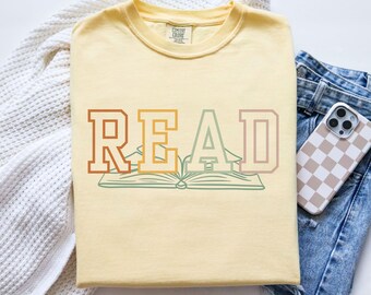 May include: A yellow t-shirt with the word "READ" in a colorful, layered font. The letters are outlined in a darker shade of the same color. A stylized open book is centered below the word.