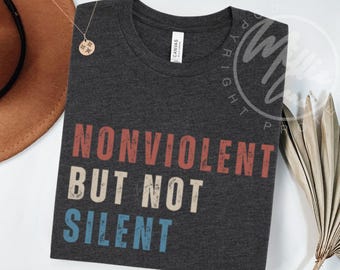 May include: Dark gray t-shirt with the words "NONVIOLENT BUT NOT SILENT" in red, white, and blue block letters. The shirt is made of a soft material and has a crew neck.