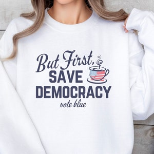 May include: White sweatshirt with the text "But First, Save Democracy vote blue" in navy blue lettering. A coffee mug with an American flag design is above the text.
