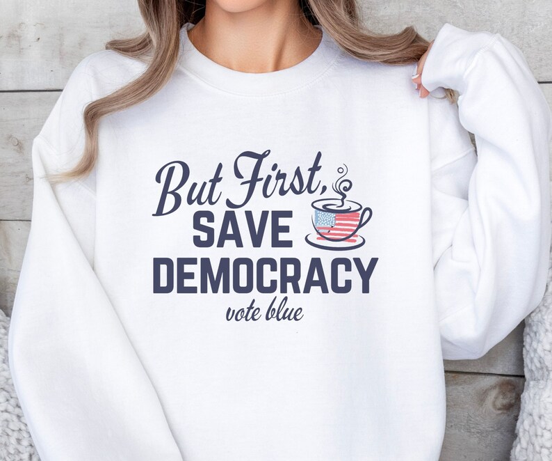 May include: White sweatshirt with the text "But First, Save Democracy vote blue" in navy blue lettering. A coffee mug with an American flag design is above the text.