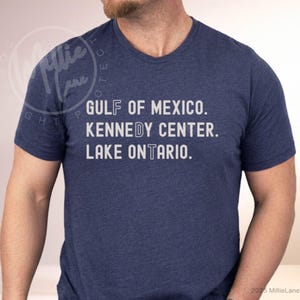 May include: A heathered navy blue short-sleeve t-shirt featuring white text that reads GULF OF MEXICO, KENNEDY CENTER, and LAKE ONTARIO. The letters F, D, and T are highlighted in a different font style within the words on the shirt.