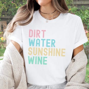 May include: White t-shirt with the words "Dirt Water Sunshine Wine" in pastel colours. The shirt has short sleeves and is made of a soft material. The text is in a playful, sans-serif font.
