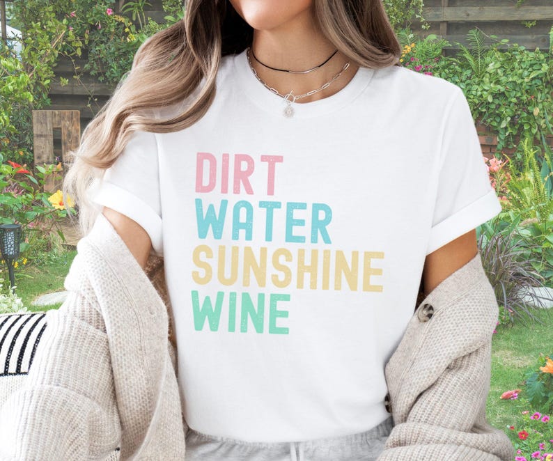 May include: White t-shirt with the words "Dirt Water Sunshine Wine" in pastel colours. The shirt has short sleeves and is made of a soft material. The text is in a playful, sans-serif font.