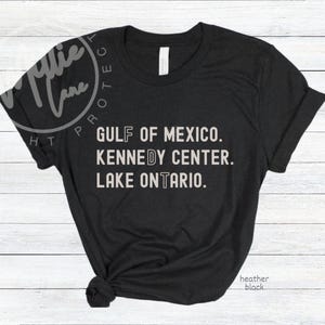 May include: A black short-sleeved t-shirt featuring white text that reads GULF OF MEXICO, KENNEDY CENTER, and LAKE ONTARIO. The letters F, D, and T are highlighted in a distinct font style. The shirt has a crew neckline and a knotted hem detail.