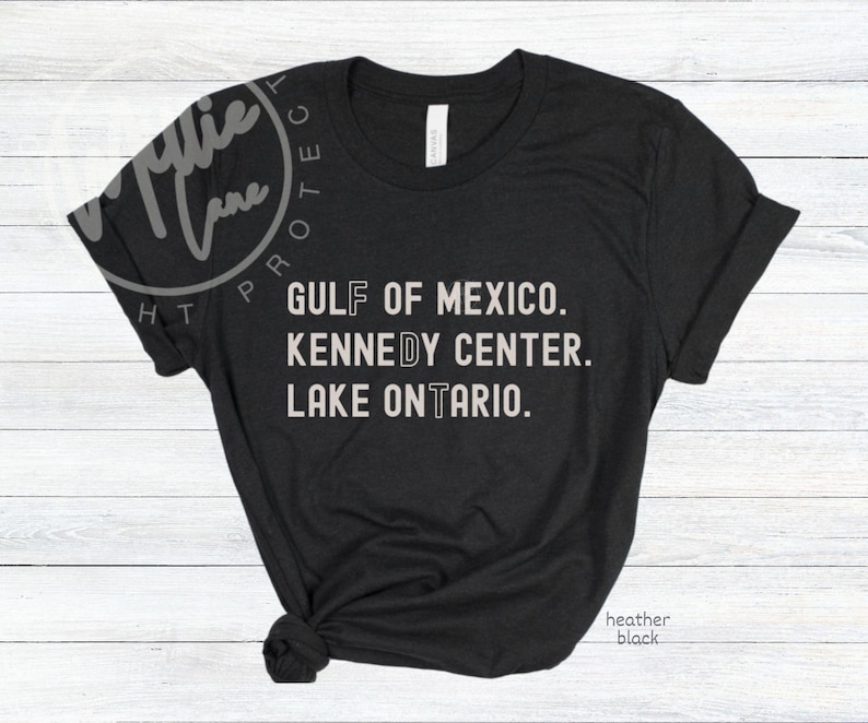 May include: A black short-sleeved t-shirt featuring white text that reads GULF OF MEXICO, KENNEDY CENTER, and LAKE ONTARIO. The letters F, D, and T are highlighted in a distinct font style. The shirt has a crew neckline and a knotted hem detail.