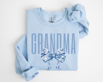 以下が含まれることがあります： 背の高いサンセリフフォントでGRANDMAという文字がプリントされた、ライトブルーのクルーネックトレーナー。文字の下には、伝統的な磁器を思わせる青と白の花柄が描かれた、大きく詳細なリボンのイラストが配置されています。