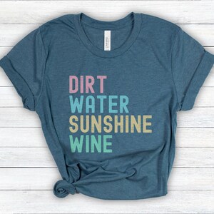 May include: A teal t-shirt featuring the words "Dirt, Water, Sunshine, Wine" in a stacked, multi-coloured font. The shirt has rolled-up sleeves and is made of a soft material. A casual wear item.