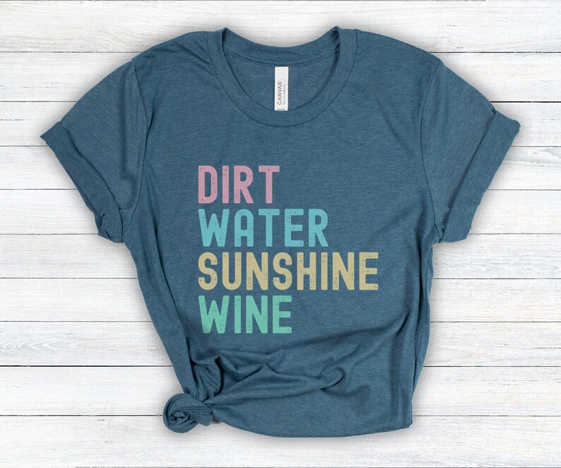 May include: A teal t-shirt featuring the words "Dirt, Water, Sunshine, Wine" in a stacked, multi-coloured font. The shirt has rolled-up sleeves and is made of a soft material. A casual wear item.